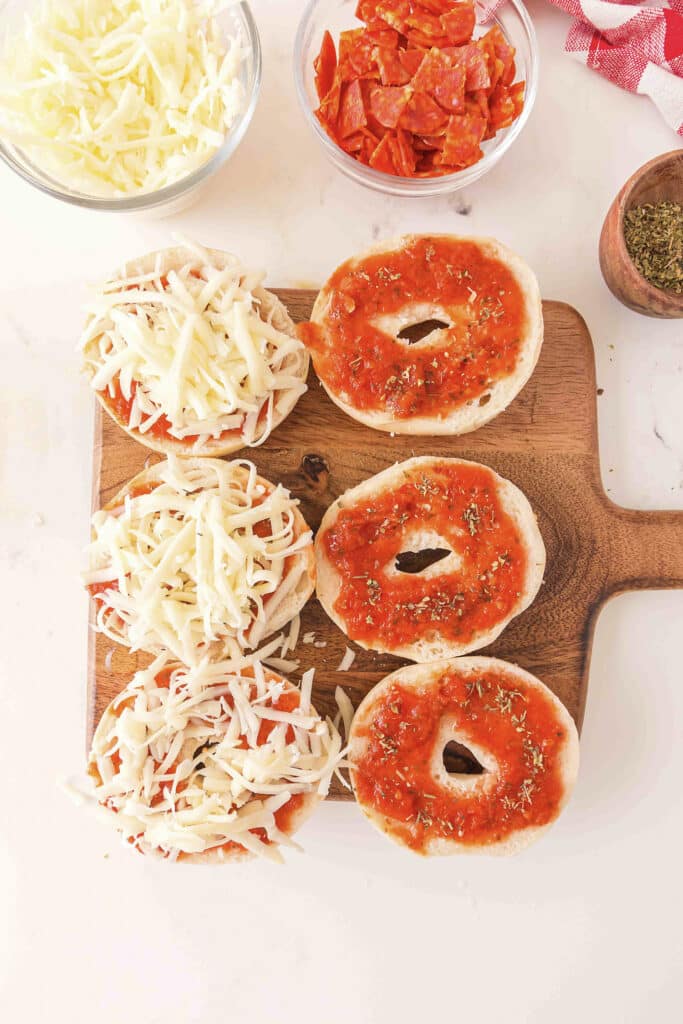 Six uncooked bagel pizzas are arranged on a wooden cutting board: three topped with sauce and cheese, and three with only pizza sauce.