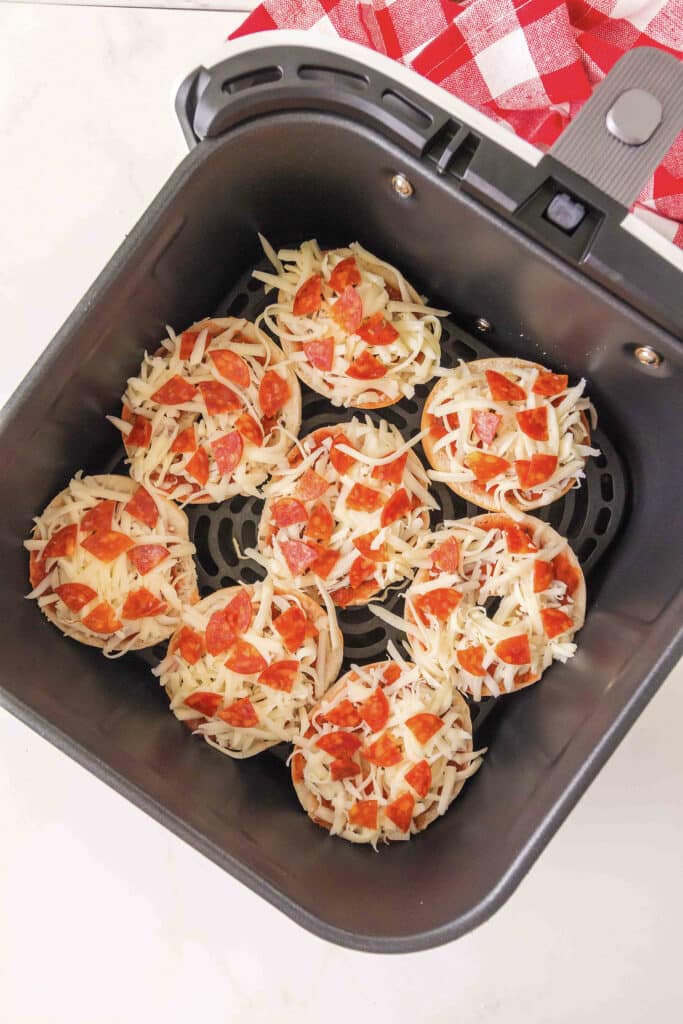 Uncooked bagel pizzas are arranged in a single layer inside an air fryer basket.