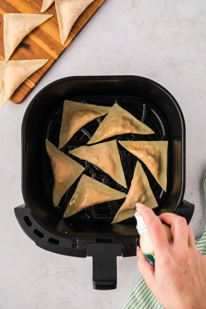 Uncooked buffalo chicken wontons are arranged in an air fryer basket while being lightly misted with oil.