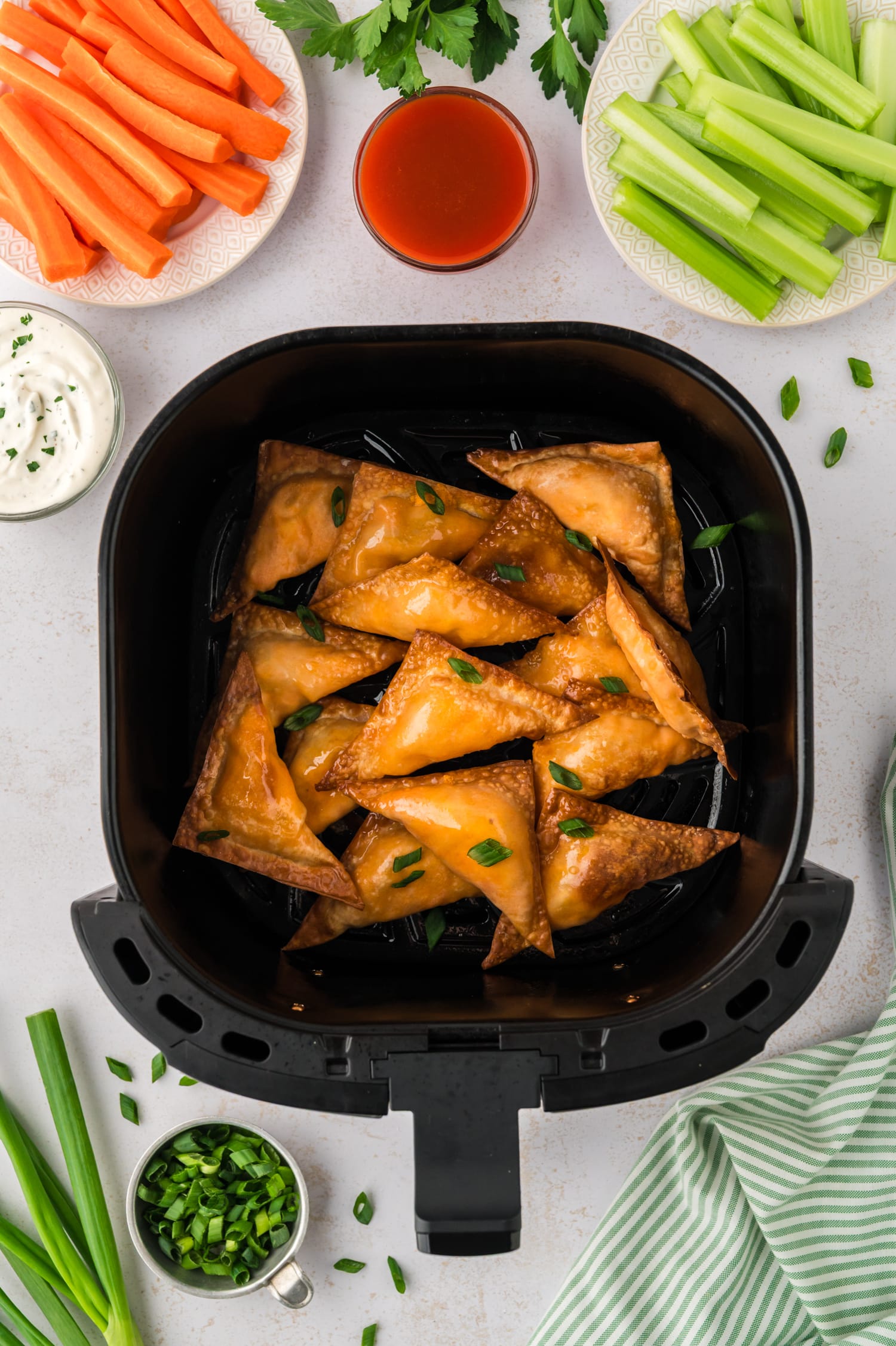 An overhead view of an air fryer basket filled with buffalo chicken wontons, with a cup of buffalo sauce positioned above the basket.