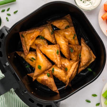 An overhead view of an air fryer basket filled with buffalo chicken wontons, placed next to separate plates of sliced carrots and celery sticks.