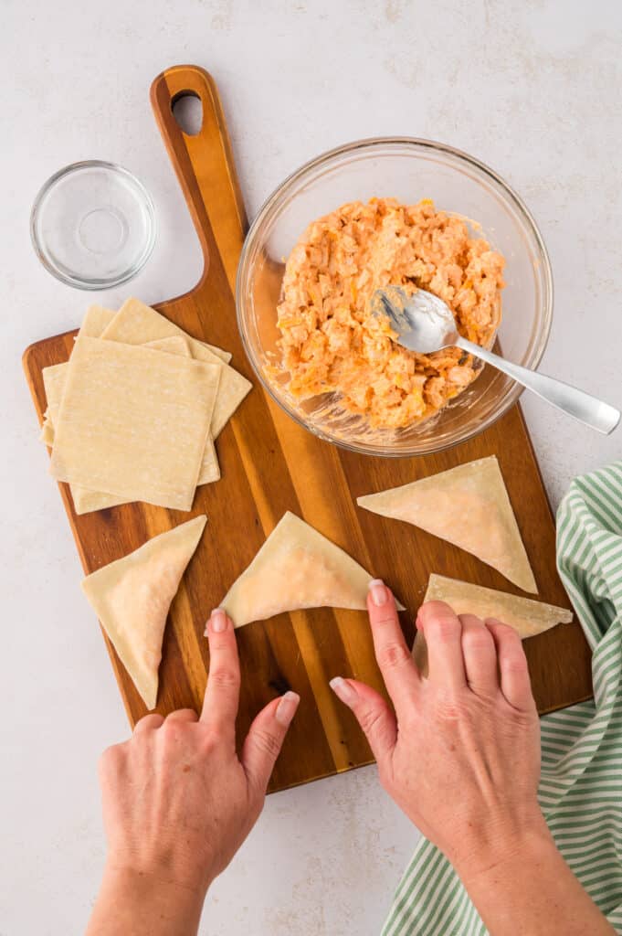 An overhead view shows a woman’s hands pressing down on the corners of a wonton to seal it.