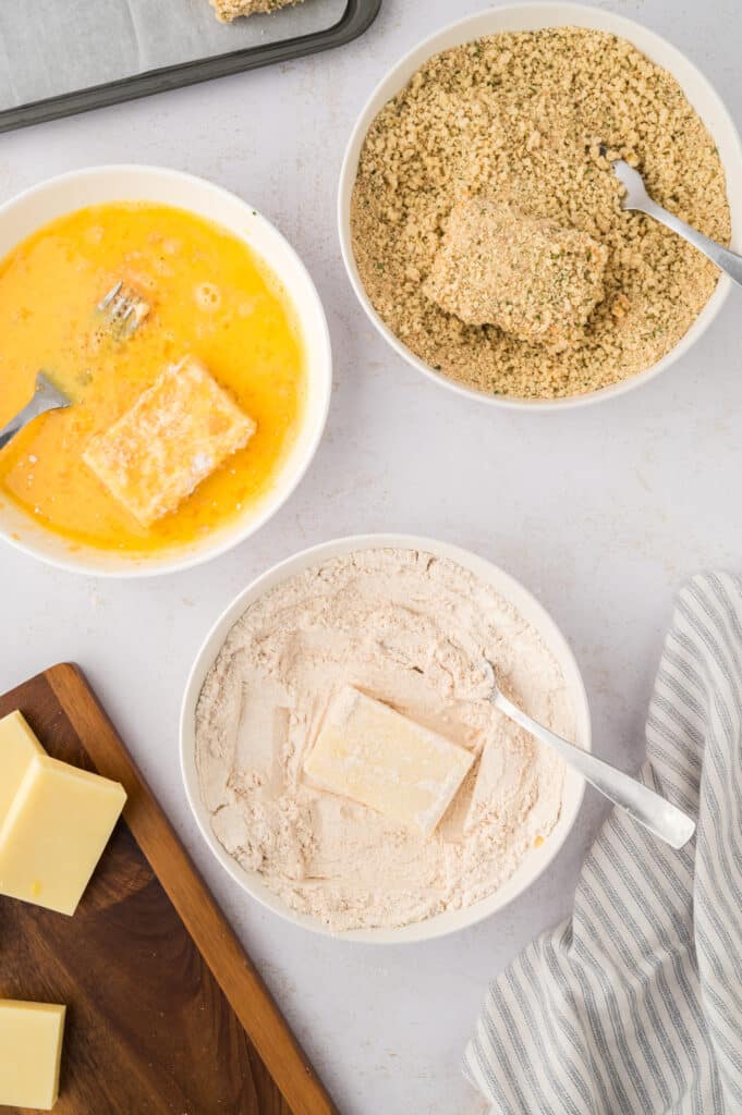 Three bowls display the steps for breading mozzarella sticks: one with flour, one with beaten egg, and one with panko breadcrumbs.
