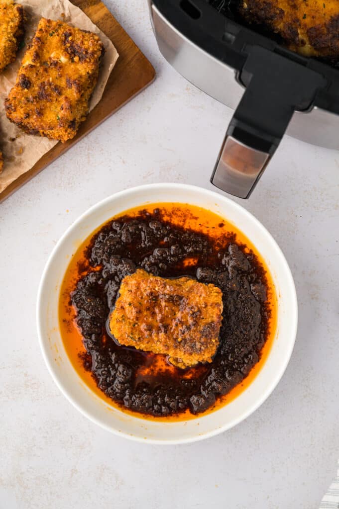 A mozzarella stick lies in a bowl of Nashville hot seasoning, its surface coated in the deep red, spicy powder.