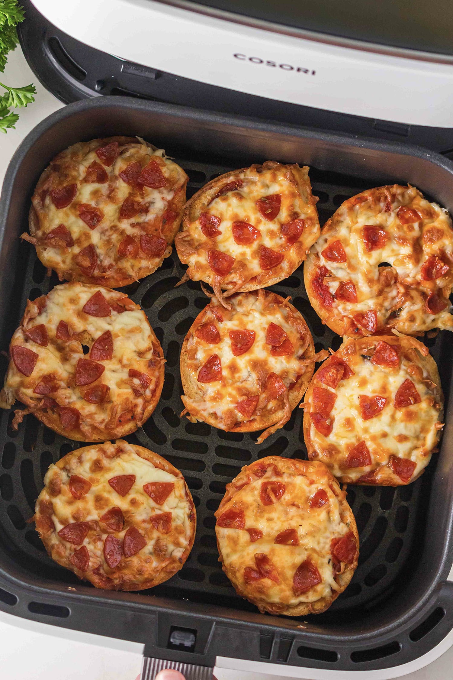 An overhead view of eight bagel pizzas arranged in a single layer inside an air fryer basket.