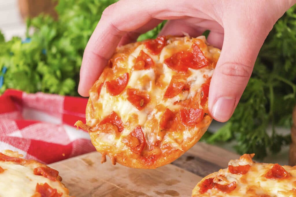 A close-up of a person holding a bagel pizza, showing the toppings and melted cheese on the surface.