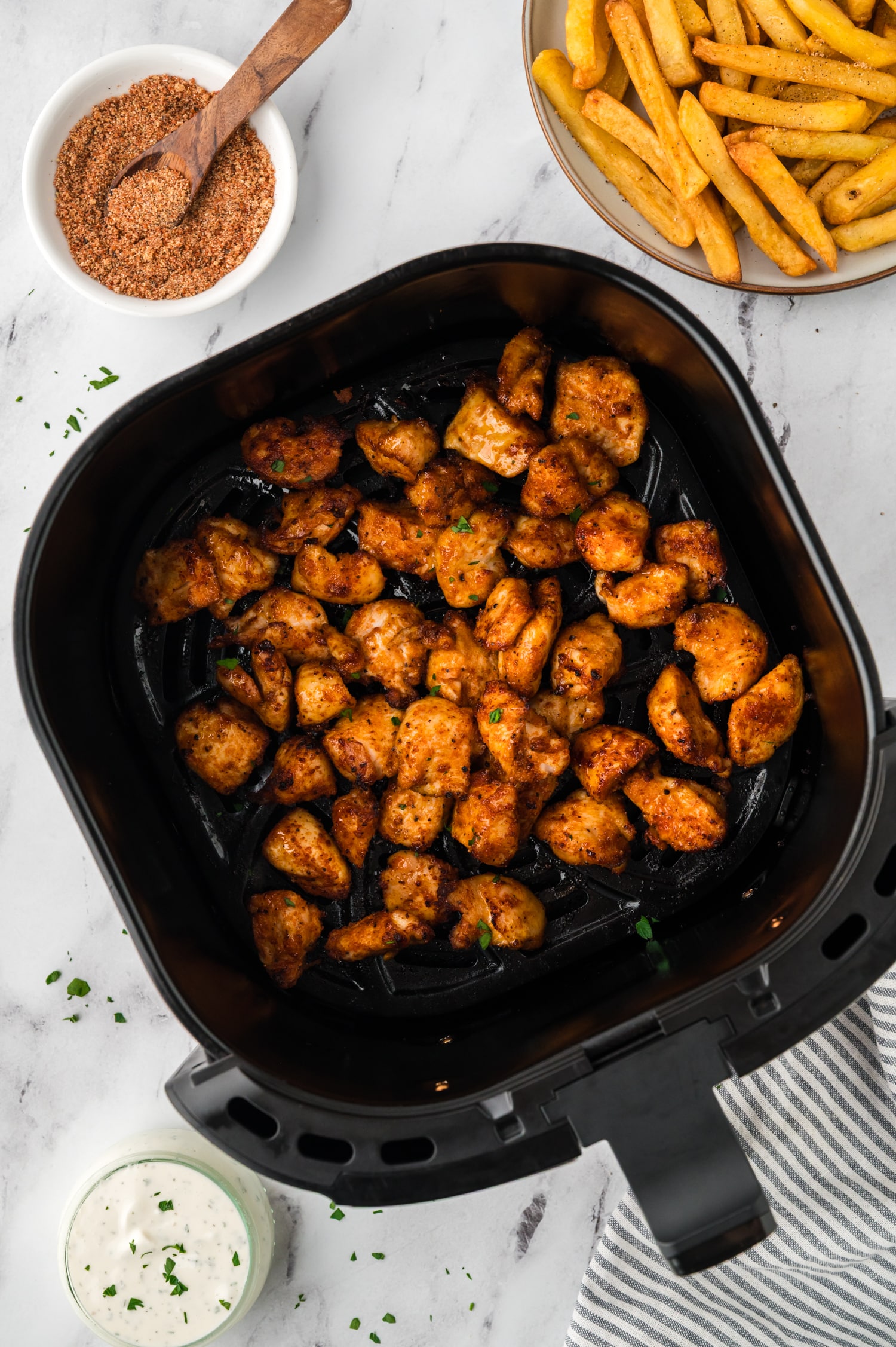 Overhead view of chicken bites in an air fryer basket next to seasoning.
