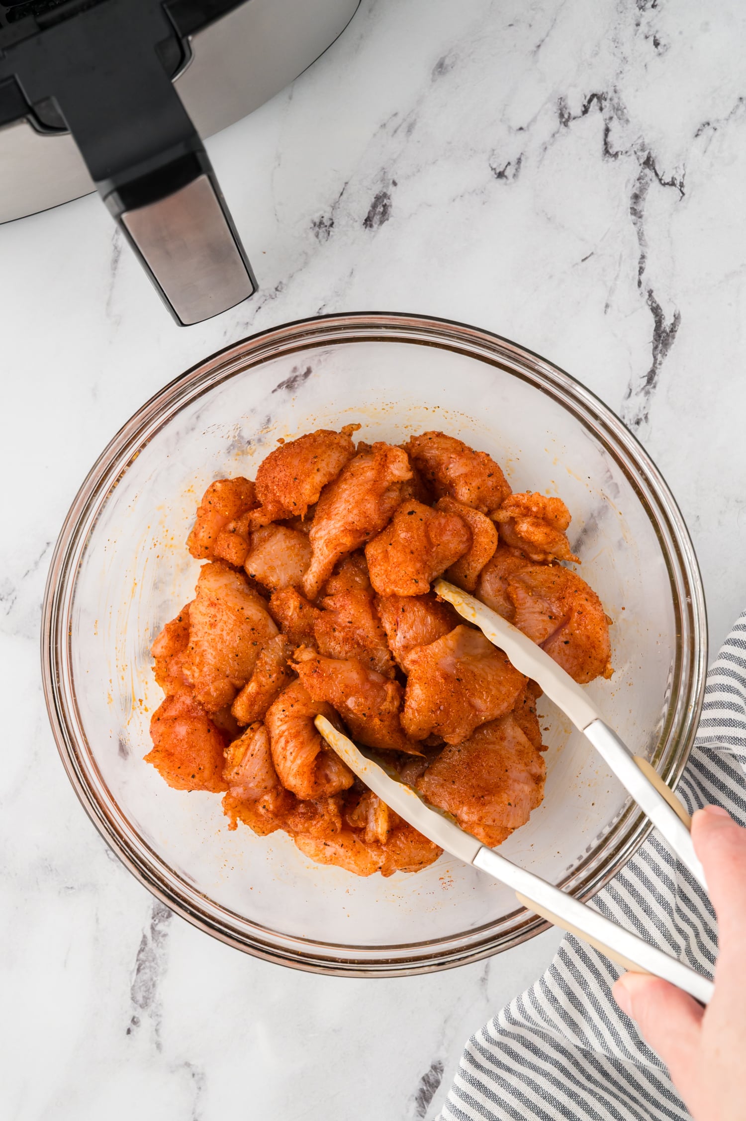 Tongs mixing raw chicken bites in a clear glass bowl.