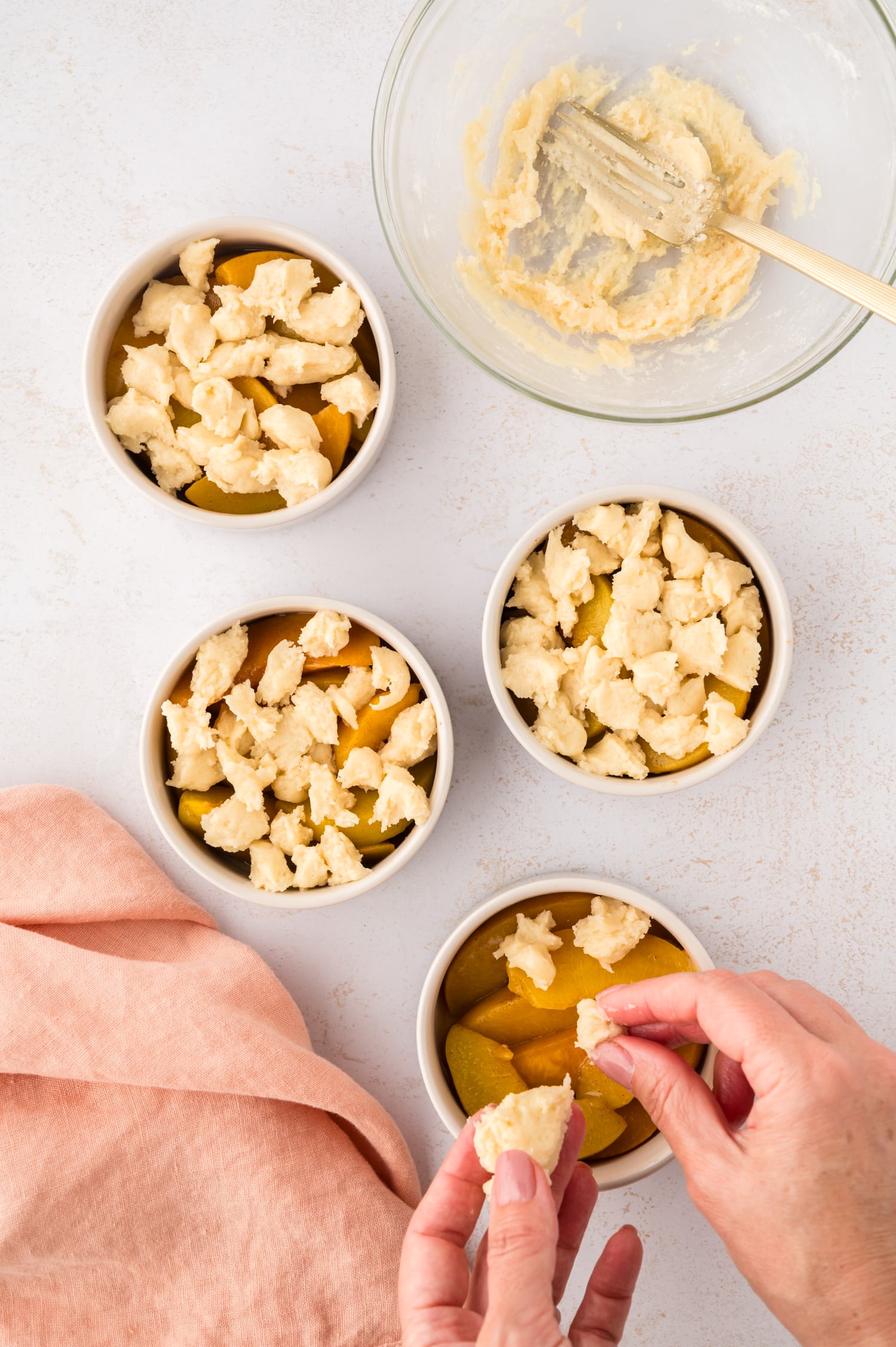 A person is breaking off pieces of raw dough and placing them on top of the seasoned peaches inside a ramekin, preparing them for baking.