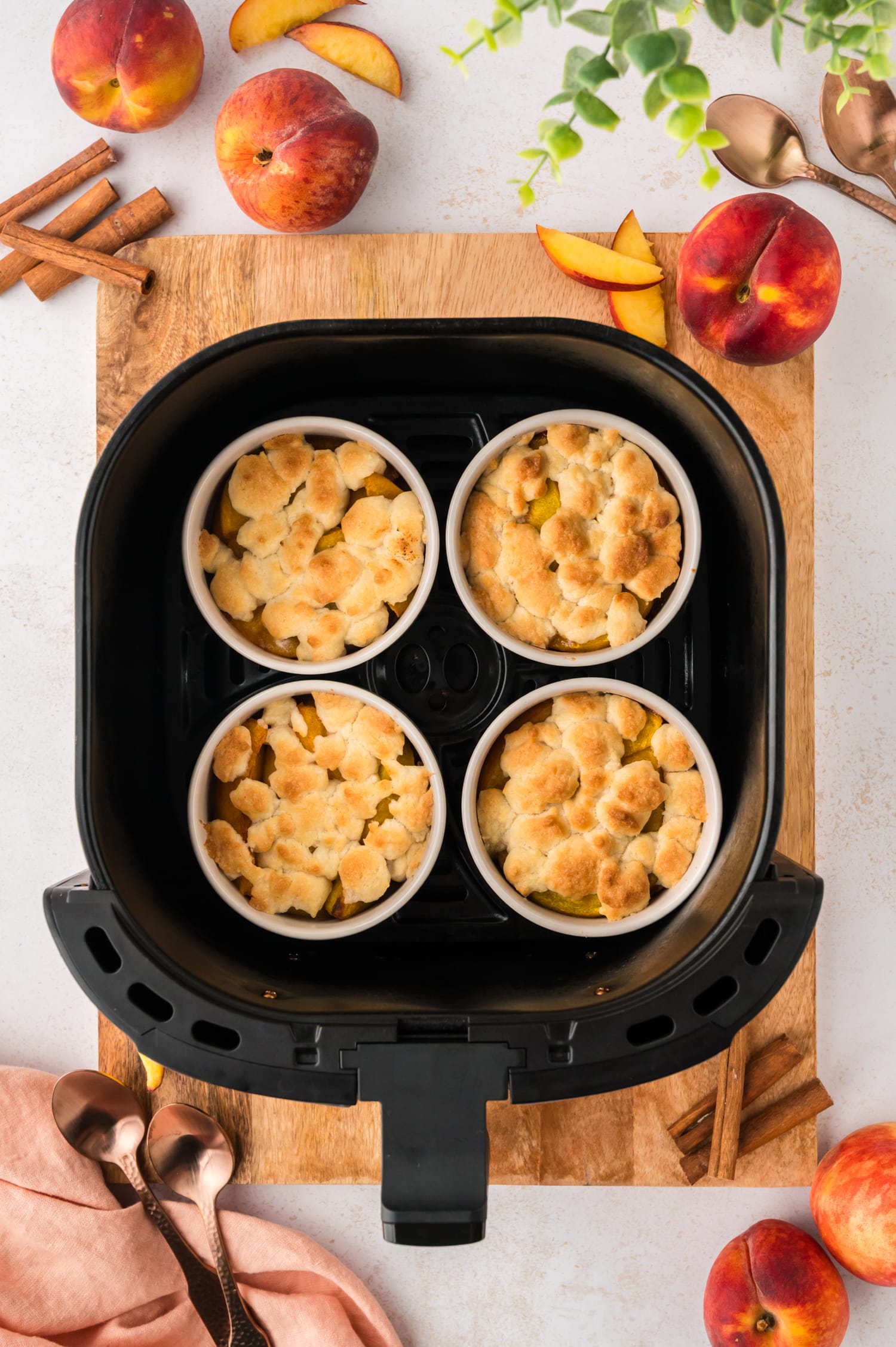 An overhead view shows an air fryer basket resting on a wooden cutting board, containing all four ramekins of mini peach cobbler inside.