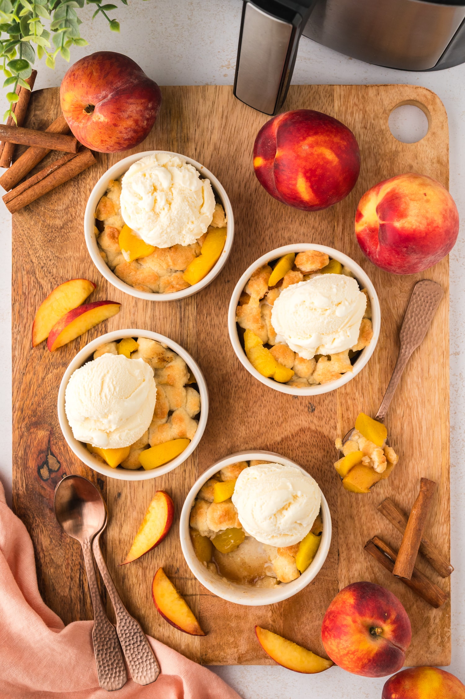 A wooden cutting board displays cinnamon sticks, On a wooden cutting board, there are cinnamon sticks, sliced peaches, whole peaches, and four ramekins of mini peach cobbler, each topped with a scoop of vanilla ice cream, all arranged together.