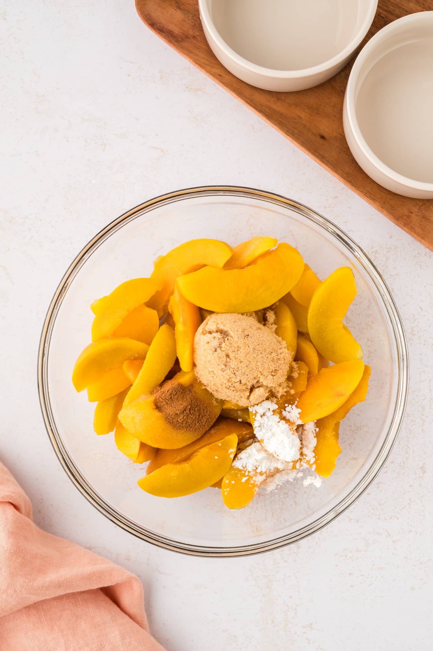 Sliced peaches are in a clear glass bowl, mixed with seasoning, preparing the cobbler filling.