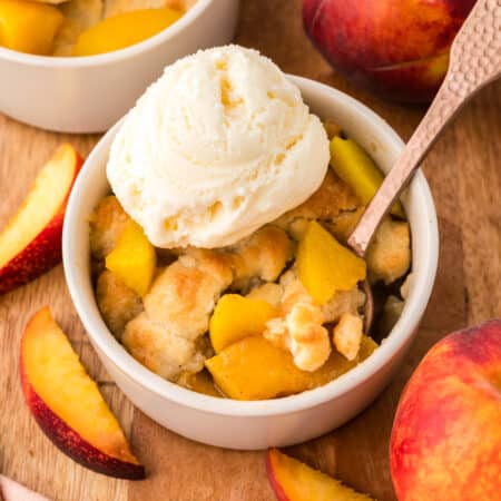 A close-up shows a mini peach cobbler with a spoon dipping A close-up shows a mini peach cobbler with a spoon dipped into it, and a scoop of vanilla ice cream resting on top of the warm cobbler.
