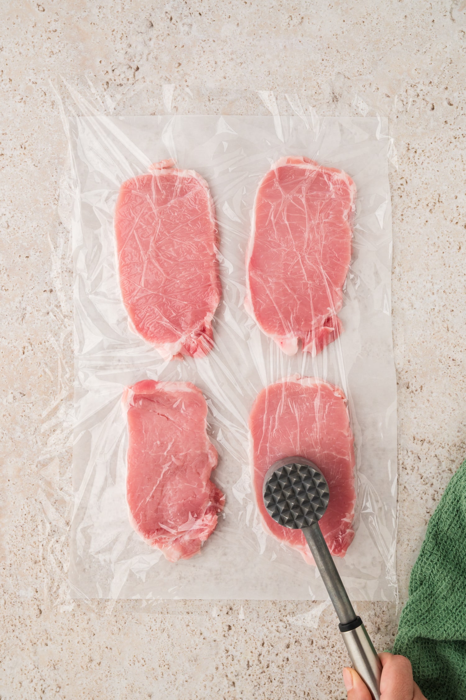 Four pork chops are placed between two pieces of parchment paper, and they’re being flattened with a meat tenderizer to even out their thickness.