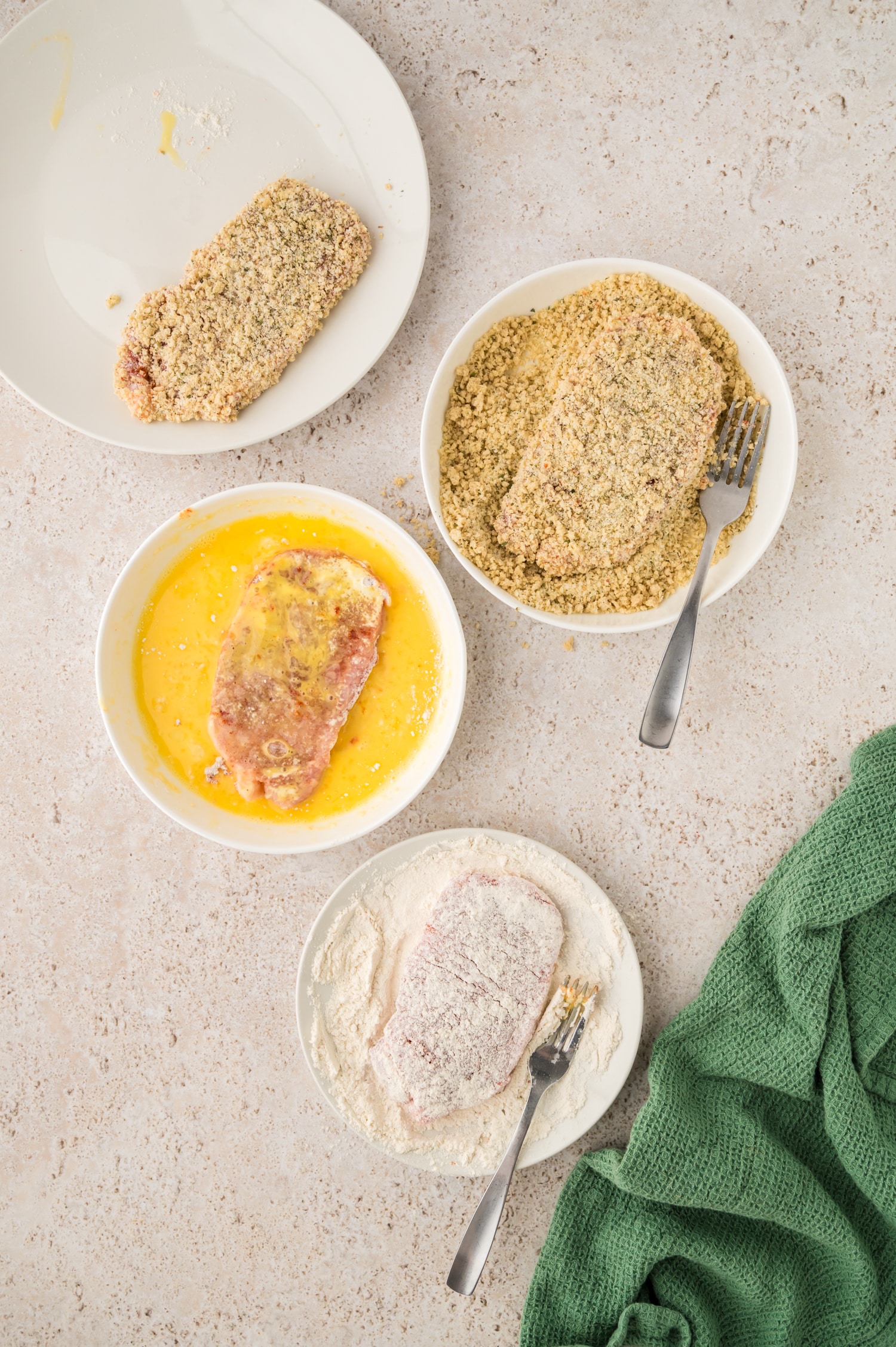 Pork milanese is being dipped in three separate bowls: one with flour, one with beaten egg, and one with breadcrumbs, preparing each piece for coating.