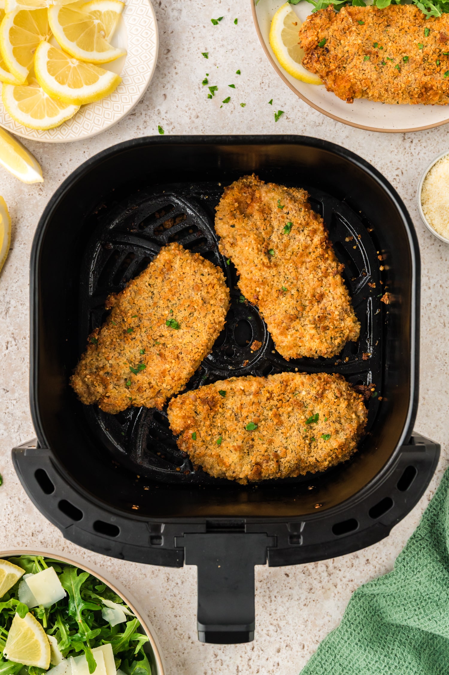An overhead view shows three pork milanese pieces inside an air fryer basket on a wet countertop.