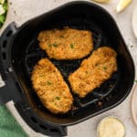 Three pork milanese pieces are inside an air fryer basket, resting on the countertop, fully cooked.