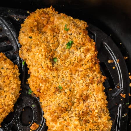 A close-up shows a single piece of pork milanese resting inside an air fryer basket, highlighting its crisp, golden-brown exterior.
