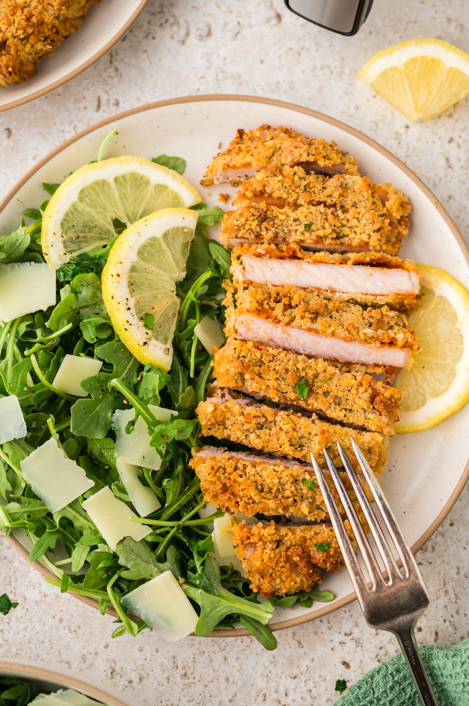 A plate holds a bed of salad greens, topped with a piece of pork milanese, and thin slices of lemon are placed on top, adding a bright finishing touch.