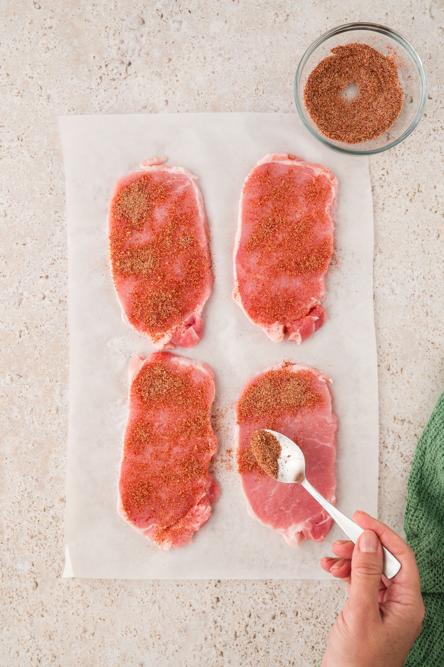 A person uses a spoon to sprinkle seasoning evenly over raw pork chops, ensuring each piece is coated before breading.