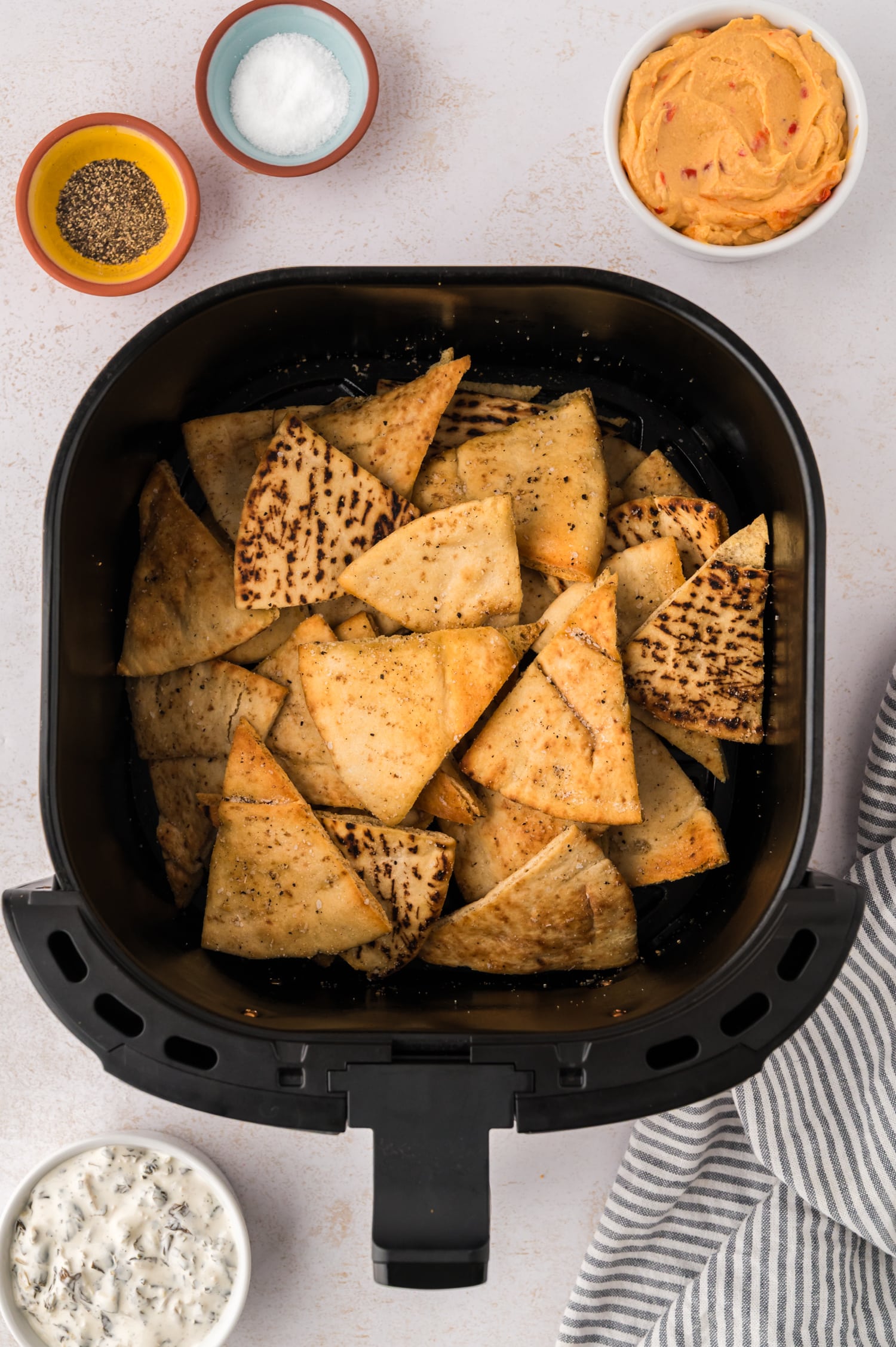 Cooked air fryer pita chips stacked in a basket, surrounded by bowls of seasonings and dips.