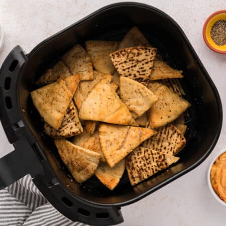 Cooked air fryer pita chips stacked in a basket, surrounded by bowls of seasonings and dips.