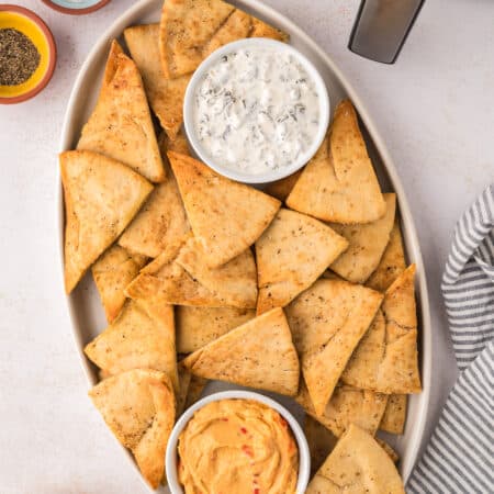 An oval bowl filled with golden-brown air fryer pita chips sits next to a metal air fryer basket.