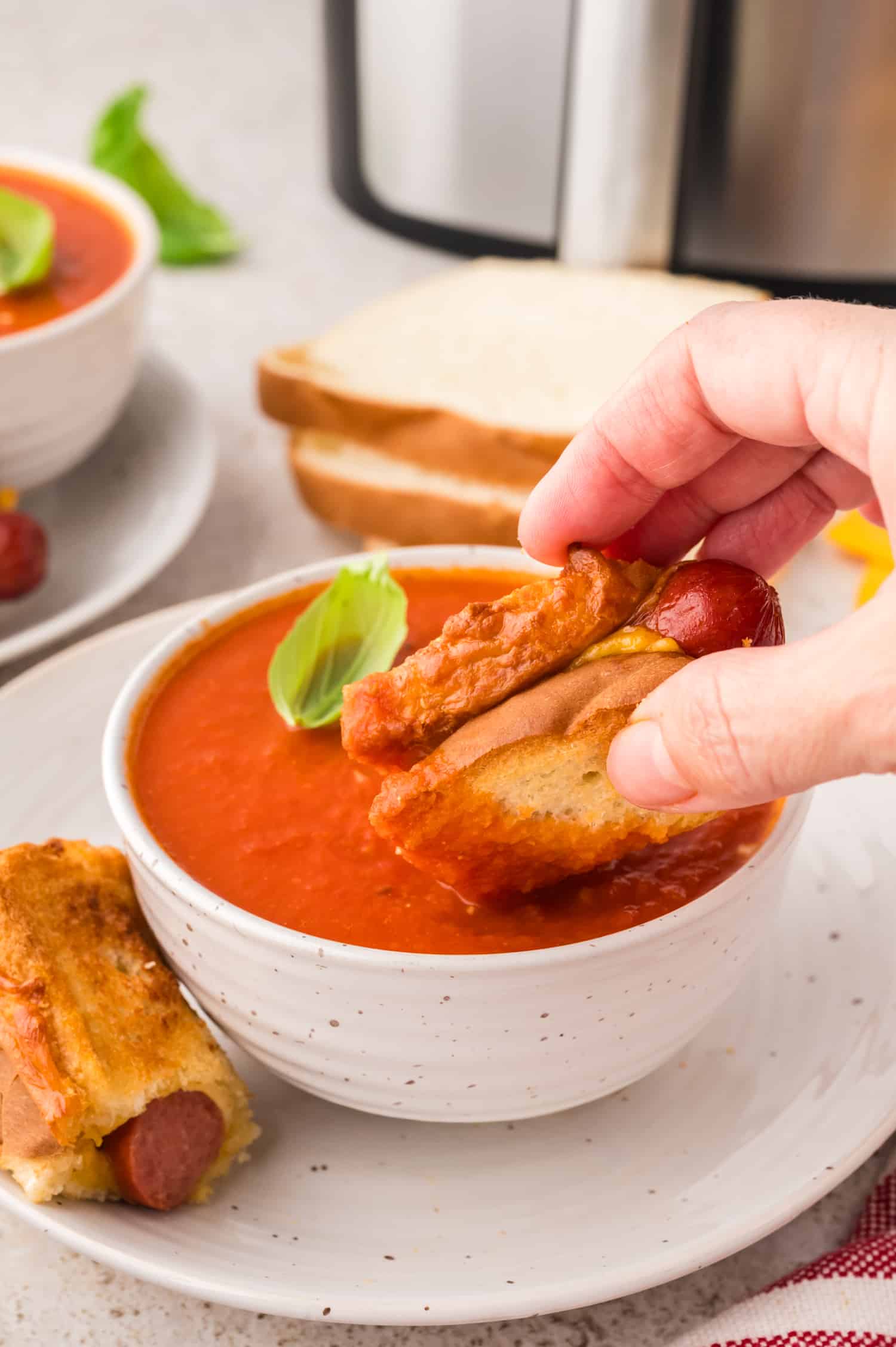 A grilled cheese hot dog being dipped into a bowl of tomato soup.