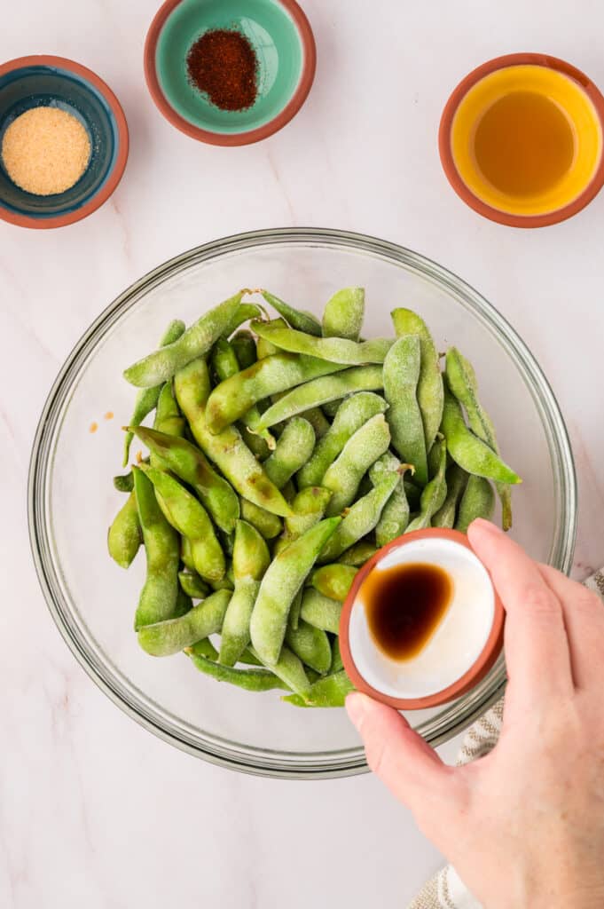 A person dumbing seasonings on raw edamame in a clear glass bowl.