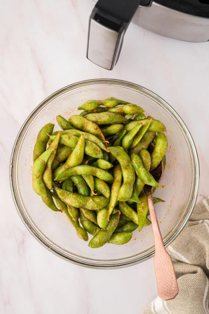 Raw edamame in a clear glass bowl with seasonings on it.