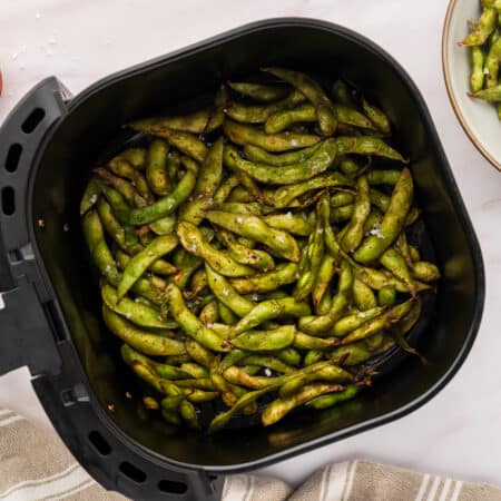 A plate of edamame next to an air fryer basket with full of it.