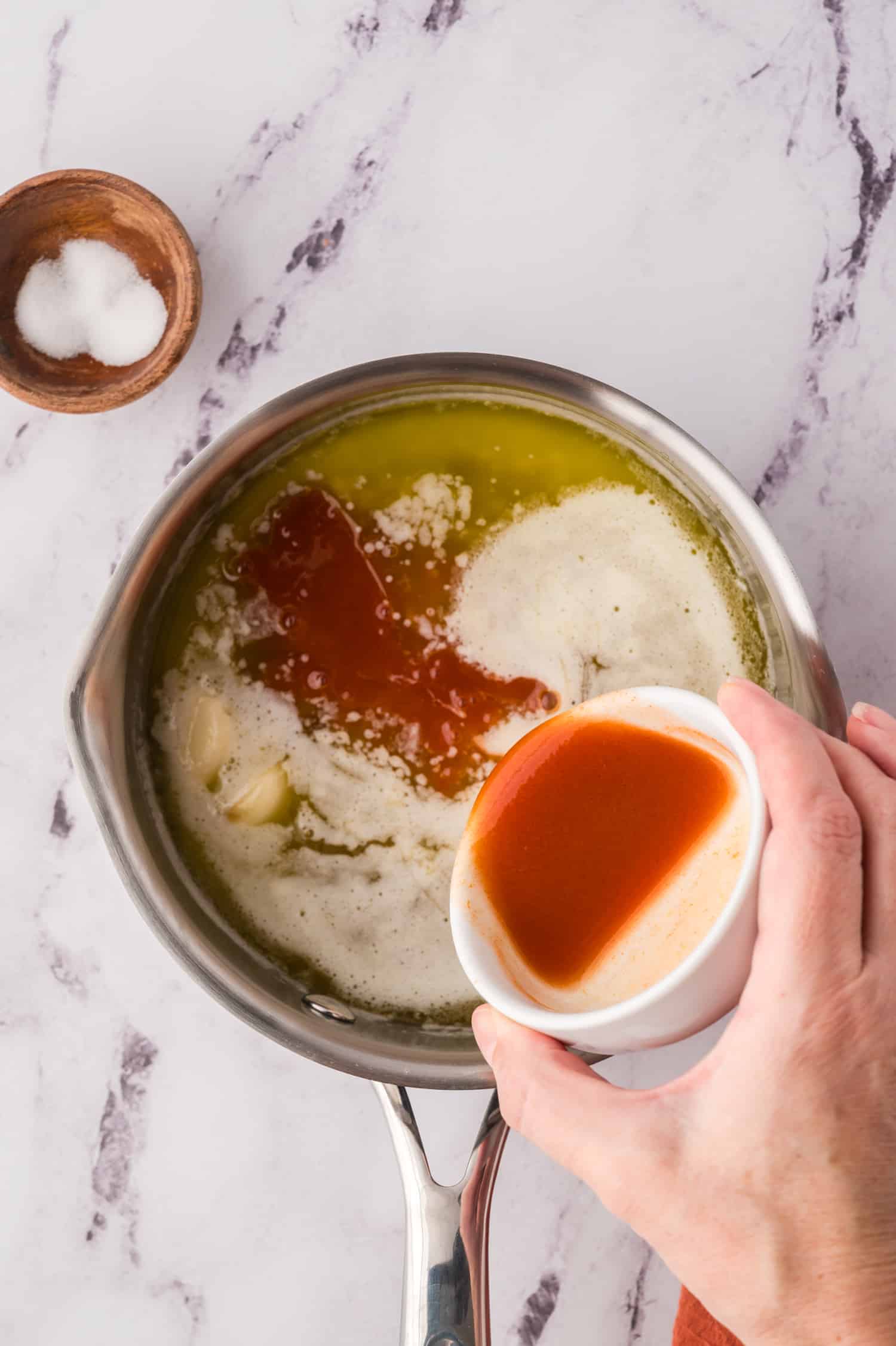 Hand pouring tabasco into sauce pan with honey.