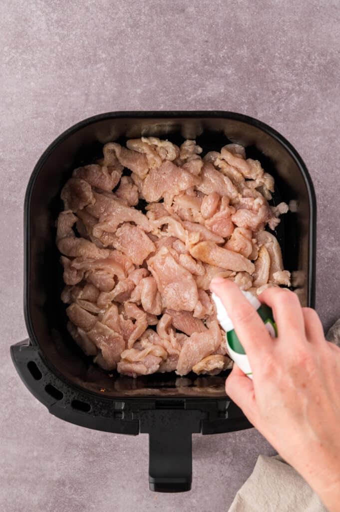 Oil being misted on raw chicken in an air fryer basket.