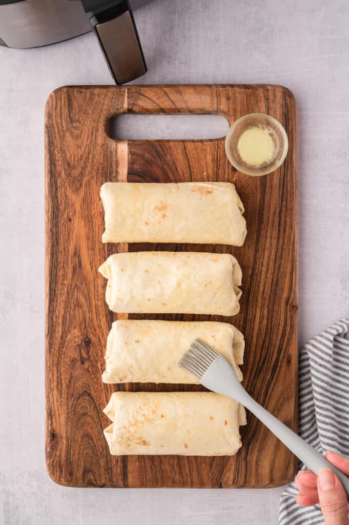 Four air fryer ham and cheese wraps on a wooden cutting board being brushed with butter.