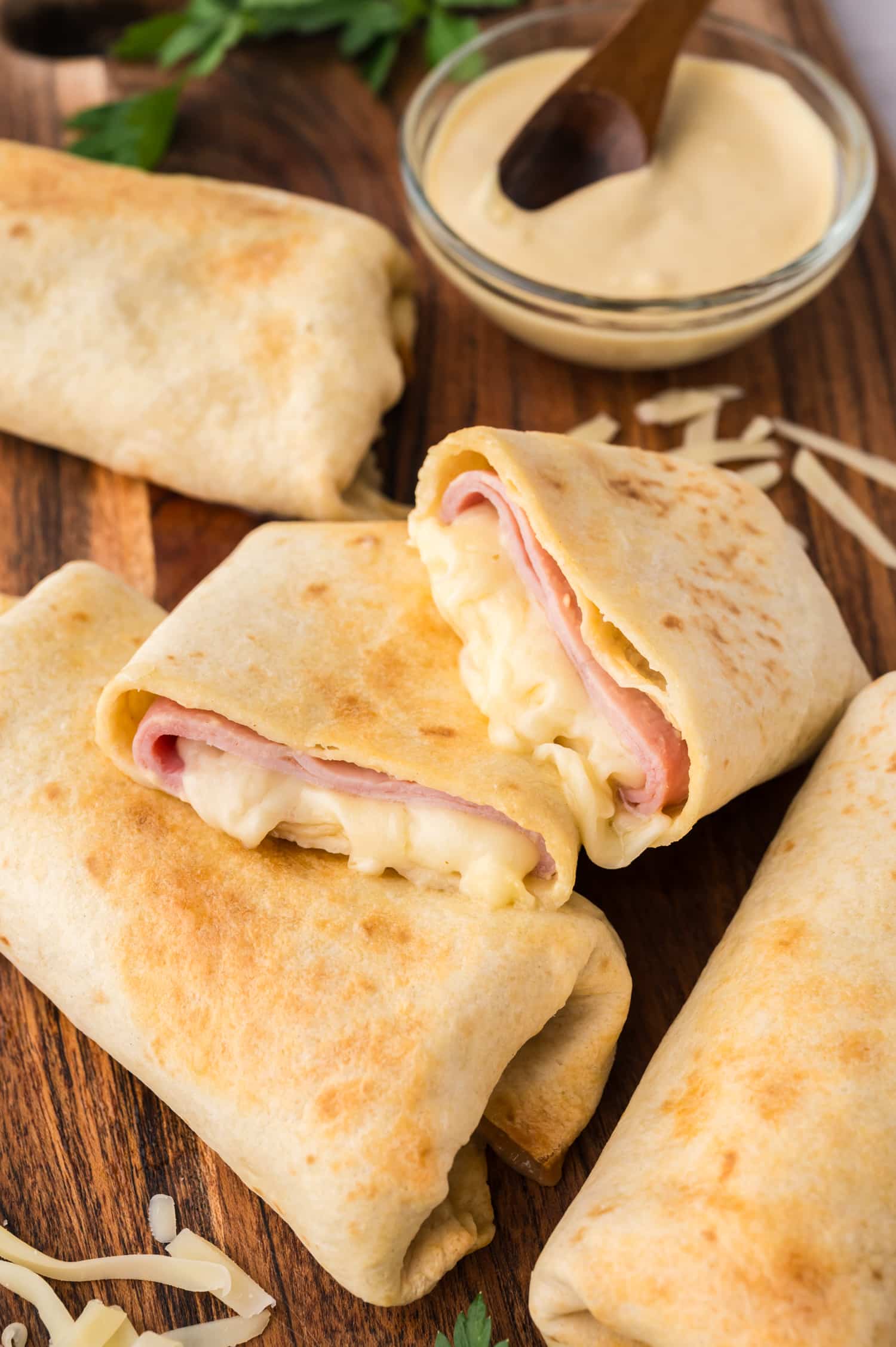 Several air fryer ham and cheese wraps on a wooden cutting board with a cheese sauce in the background.
