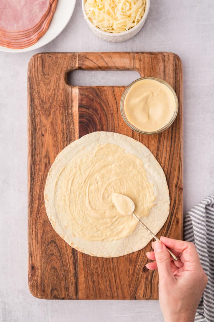 A spoon spreading sauce on a tortilla in preparation for an air fryer ham and cheese wrap.